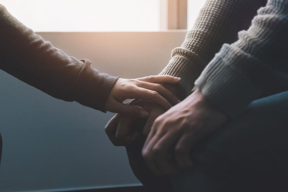 Two people sitting by a window, one clasping the other's hands in support.