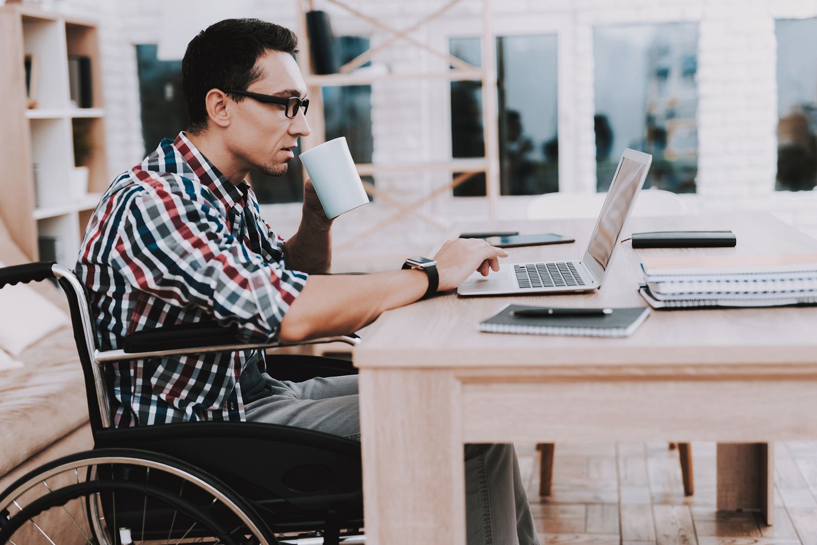 A man using a wheelchair works on a laptop at a table at home, drinking coffee.